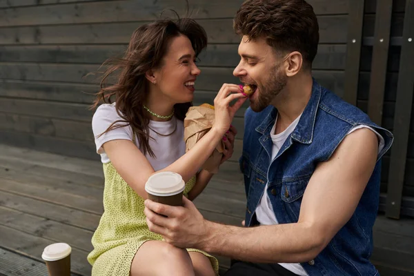 stock image Cheerful and stylish brunette woman in sundress feeding boyfriend with bun and sitting near coffee to go and wooden house at background, serene ambiance concept, tranquility
