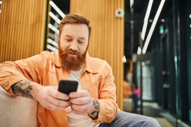 Sakallı ve dövmeli bir iş adamı cep telefonuyla iletişim kurarken aynı zamanda modern çalışma odasının bulanık arka planında rahat bir kanepede oturuyor.
