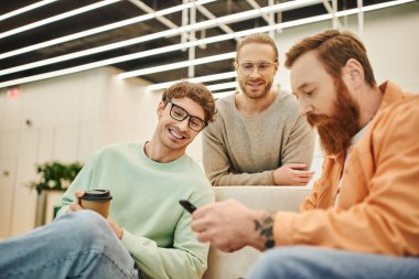 cheerful businessman with takeaway drink looking at bearded colleague networking on mobile phone on couch in lounge of modern office environment during coffee break, blurred foreground clipart