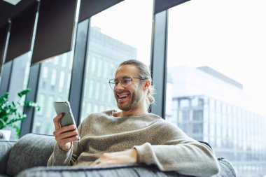 Heyecanlı ve şık gözlüklü iş adamı ve güncel giysiler içinde modern ofis salonunda büyük pencerelerin yanında oturuyor ve cep telefonlarına, iş ilhamlarına ve pozitifliğe bakıyor.