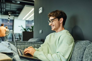Modern ofis, iş ilhamı ve verimliliğinde rahat bir koltukta otururken gündelik giysiler içinde neşeli iş adamı ve dizüstü bilgisayarda yazı yazan şık gözlükler.