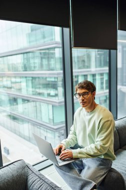 ambitious and career oriented businessman in stylish eyeglasses and casual clothes sitting on comfortable couch with laptop and looking at camera near large windows in lounge of modern office clipart