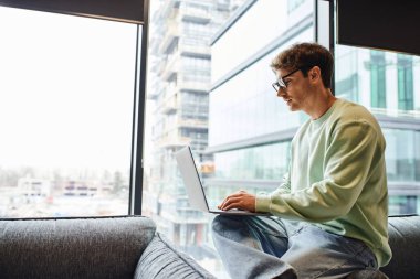 Modern ofis alanındaki büyük pencerelerin yanında oturan ve dizüstü bilgisayar, iş ilhamı ve hırs konsepti üzerine yeni bir başlangıç planı üzerinde çalışan sıradan giyinmiş ve gözlüklü bir adamın yan görüntüsü.