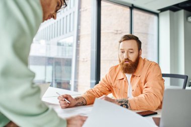 bearded architectural designer holding pen and working with blueprint near smartphone, laptop and colleague on blurred foreground in modern coworking office, business and creativity concept clipart