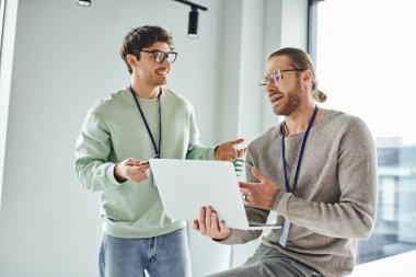 optimistic designer in eyeglasses discussing startup project with colleague holding laptop in modern architectural studio, concept of creativity and successful business collaboration clipart