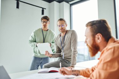 ciddi mimarlar gözlüklü ve gündelik giysiler içinde meslektaşlarının yanında dizüstü bilgisayara bakıyorlar modern tasarım stüdyosunun çalışma ofisinde bulanık önplan üzerinde çalışıyorlar.