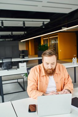 focused, bearded and tattooed businessman looking at laptop while sitting at modern workplace near smartphone with blank screen in contemporary office space and working on startup project clipart