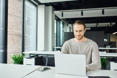 smiling entrepreneur in stylish eyeglasses planning new business project and working on laptop near mobile phone with blank screen on work desk in modern coworking environment  clipart