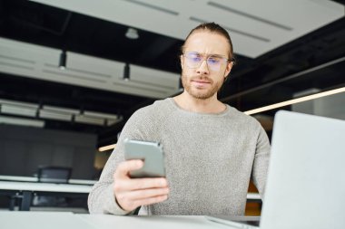 Gözlüklü ve günlük kıyafetli girişimci, ciddi yüz ifadesi olan, modern ofis alanındaki bulanık ön planda dizüstü bilgisayar projesinde çalışırken cep telefonuna bakan