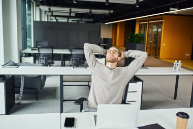 Yorgun bir iş adamı, elleri ve gözleri kapalı, iş yerinde dizüstü bilgisayarın yanında otururken, modern ofis ortamında boş ekranlı, akıllı bir telefonla dinleniyor.