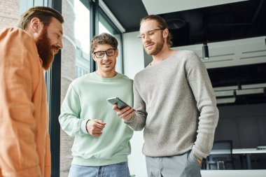 Gözlüklü ve şık giyimli iş adamı cep telefonu ve cep telefonuyla çağdaş ofisteki gülümseyen iş arkadaşlarının yanında duruyor, başarılı işbirliği kavramı.
