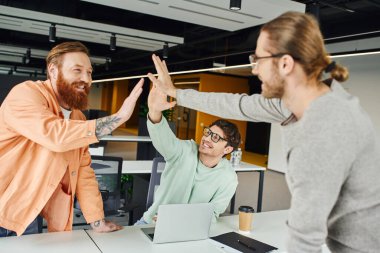 excited business team, bearded tattooed man and colleagues in eyeglasses giving high five while confirming agreement near laptop in modern coworking office, concept of successful collaboration clipart