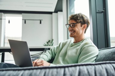 Şık gözlükleri ve günlük kıyafetleriyle neşeli ve başarılı iş adamı çalışma odasındaki rahat koltukta oturuyor ve laptopta iletişim kuruyor, iş ilhamı kavramı