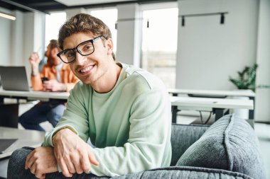 happy entrepreneur in stylish eyeglasses and casual clothes sitting in armchair and smiling at camera near colleague working on blurred background in modern office, productive coworking concept clipart