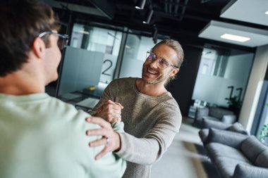 excited and satisfied entrepreneur in eyeglasses shaking hands and touching shoulder while closing deal with business partner in modern lounge of coworking office, successful collaboration concept clipart