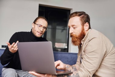 businessman in black turtleneck and eyeglasses gesturing while talking to bearded tattooed entrepreneur showing startup project on laptop in modern office, business partnership concept clipart