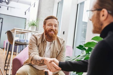 overjoyed, tattooed and bearded entrepreneur in casual clothes confirming agreement and shaking hands with businessman on blurred foreground, successful partnership concept clipart