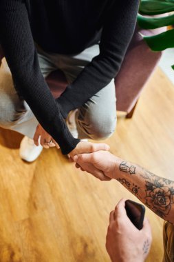 top view of entrepreneur in black turtleneck shaking hands with tattooed businessman with mobile phone, cropped view, successful partnership concept clipart