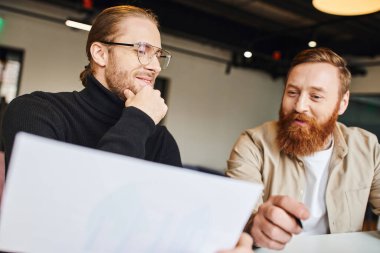 Pleased businessman in black turtleneck and eyeglasses touching chin while looking at document near happy bearded colleague while working on new startup in coworking office stock vector