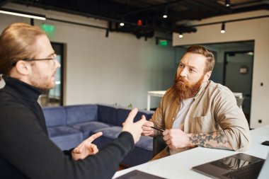 attentive, bearded and tattooed entrepreneur holding pen and listening to business partner in black turtleneck talking about startup project near laptop in contemporary office environment clipart