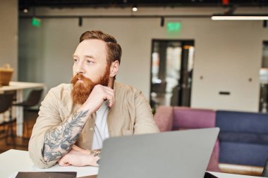 Ciddi, sakallı ve dövmeli iş adamının profesyonel portresi dizüstü bilgisayarın yanında otururken ve modern ofis ortamına bakarken, iş hayatı konsepti