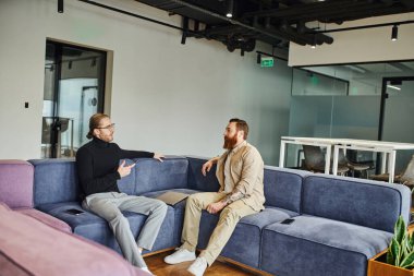 Siyah balıkçı yaka ve gözlüklü işadamı, modern ofis, ortaklık ve başarı konseptinde yumuşak koltukta sakallı meslektaşıyla konuşurken parmağıyla işaret ediyor.