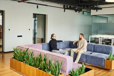 serious businessmen in stylish casual clothes sitting on soft couch near green plants and discussing startup project in modern office lounge with high tech interior, partnership and success concept clipart