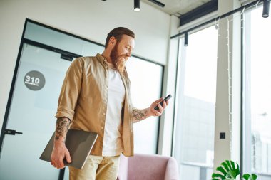 Memnun, sakallı ve dövmeli bir iş adamı cep telefonuyla internete göz gezdirirken modern iş yerinde dizüstü bilgisayarla ayakta duruyor, başarılı girişimcilik kavramı.