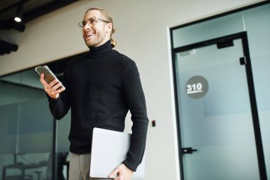 şık ve hırslı gözlüklü işadamı ve siyah balıkçı yakalı akıllı telefon ve dizüstü bilgisayarlı modern ofiste gülümseyen ve başka yöne bakan başarılı bir iş adamı.