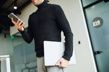 Siyahi balıkçı yakalı girişimcinin elinde dizüstü bilgisayar ve cep telefonuyla modern çalışma ofisinde dururken cep telefonuyla iletişim kurması, başarılı iş konsepti