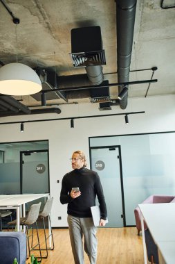 Kariyer odaklı siyah balıkçı yaka ve şık gözlükler takan iş adamı modern çalışma alanına bakarken dizüstü bilgisayar ve cep telefonuyla yürüyor, başarılı bir iş konsepti.