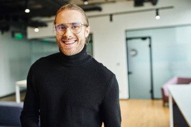 Modern ofis ortamında kameraya gülümseyen şık gözlüklü ve siyah balıkçı yakalı neşeli girişimcinin profesyonel portresi, başarılı iş konsepti