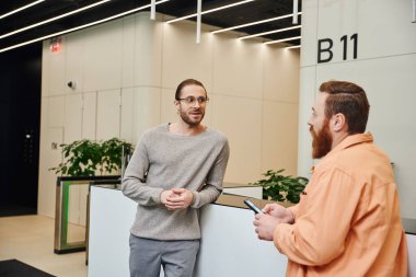 excited businessman in eyeglasses and casual clothes talking to bearded colleague at reception in lobby of contemporary coworking office, entrepreneurs discussing possibility of startup clipart
