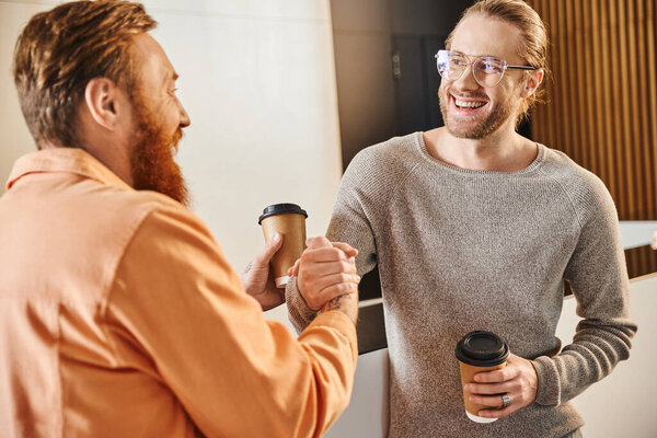 бородатый бизнесмен пожимает руку с восторженным коллегой в очках в современном офисе, успешные предприниматели с бумажными стаканчиками заключают договор на запуск во время кофе-брейка