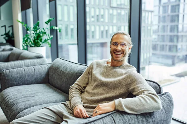 stock image happy entrepreneur in stylish eyeglasses and casual clothes, with radiant smile, sitting on comfortable couch near large windows in office lounge and looking at camera, business success concept