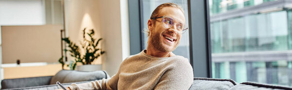 stylish and optimistic businessman in eyeglasses and casual clothes sitting in lounge of modern coworking office, smiling and looking away through window, business success concept, banner