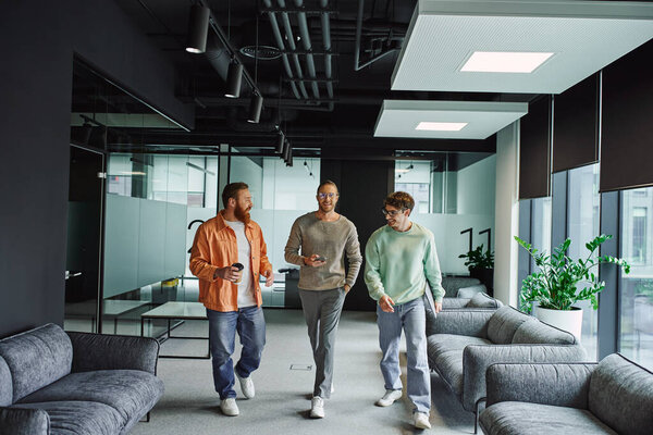 optimistic businessmen in stylish casual clothes, with paper cup, smartphone and laptop, walking and talking in modern office lounge with comfortable couches, startup discussing and collaboration