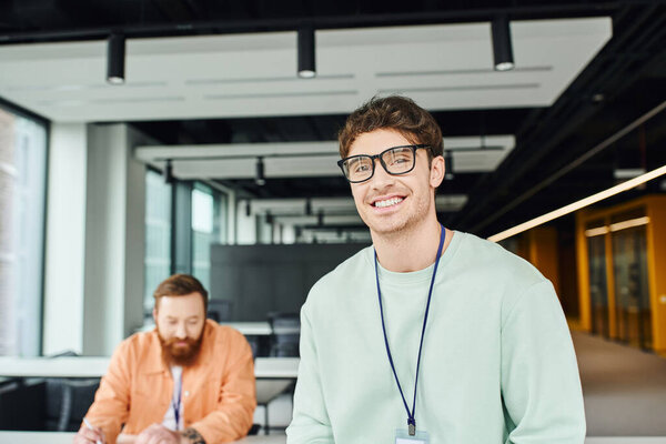 cheerful and optimistic architectural designer in eyeglasses smiling at camera near bearded colleague working on business startup on blurred background in modern office environment