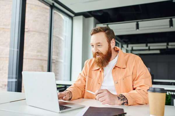 joyful, bearded and tattooed entrepreneur in casual clothes looking at laptop while working on business project near folder and paper cup with takeaway drink in contemporary office space