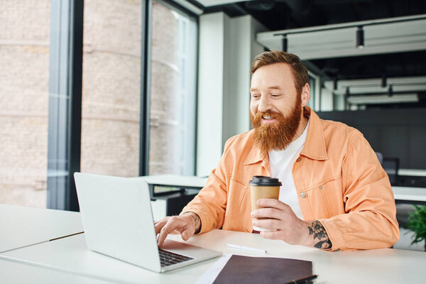 cheerful, tattooed and bearded entrepreneur holding paper cup with coffee to go and networking on laptop near folder on work desk in contemporary coworking office, successful business concept