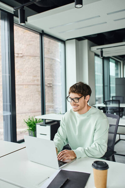 cheerful entrepreneur in stylish eyeglasses and casual clothes sitting at modern workplace and networking on laptop near folder and coffee to go in contemporary office, successful business concept