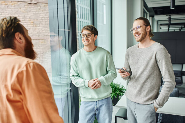 smiling businessmen in eyeglasses and casual clothes listening to bearded team lead talking on blurred foreground near large windows in contemporary office, concept of successful collaboration