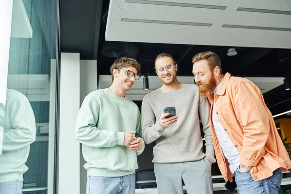 positive team lead in stylish casual clothes holding hand in pocket and browsing internet on smartphone near smiling colleagues in contemporary coworking office, concept of successful collaboration
