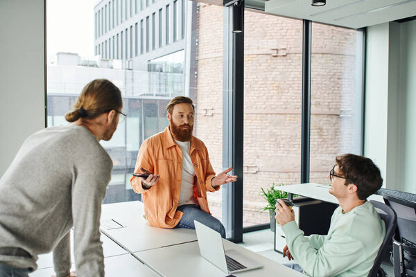 bearded team lead with mobile phone talking and discussing startup project with colleagues near laptop in contemporary coworking environment, concept of successful collaboration