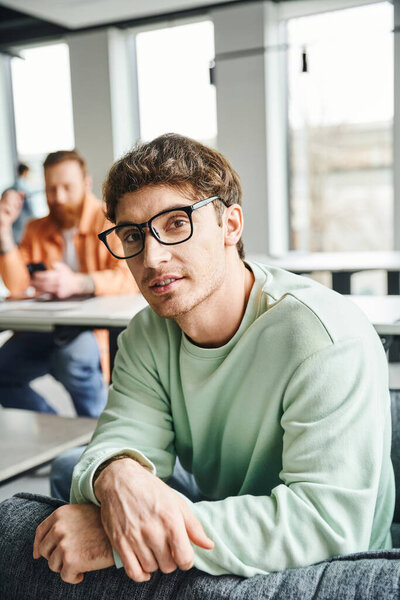 positive and ambitious entrepreneur in eyeglasses and casual clothes looking at camera while his business colleague working on blurred background in contemporary office, productive coworking concept