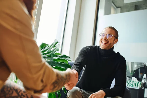 excited businessman in eyeglasses and black turtleneck shaking hands with tattooed businessman on blurred foreground in modern office, successful partnership concept