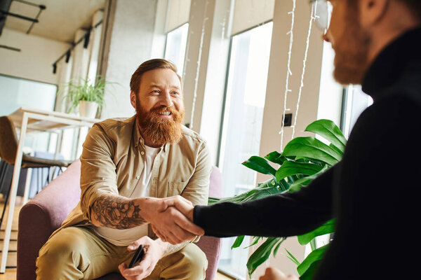 веселый, бородатый и татуированный бизнесмен пожимает руку амбициозному предпринимателю в черной водолазке, сидя в современном офисе, успешная концепция партнерства