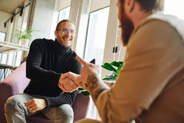 happy entrepreneur in eyeglasses and black turtleneck shaking hands and closing deal with tattooed businessman pointing with finger on blurred foreground, successful partnership concept