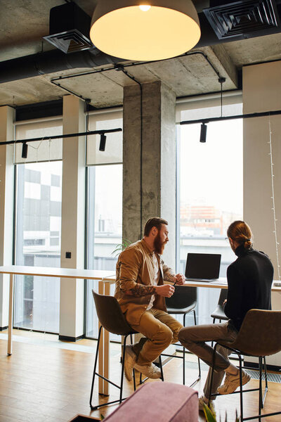 bearded and tattooed entrepreneur with business colleague in black turtleneck sitting near laptop with blank screen while working on new project in contemporary office with high tech interior 