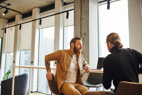 serious and bearded businessman attentively listening to colleague during startup discussing in modern office with large windows and high tech interior, productivity and collaboration concept